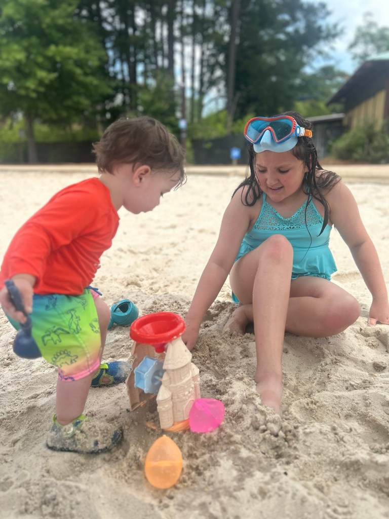 A toddler in a red long-sleeve shirt and colorful swim trunks playing in the sand with a blue toy, while a girl in a swimsuit assists in building a sandcastle with colorful molds and a red bucket.