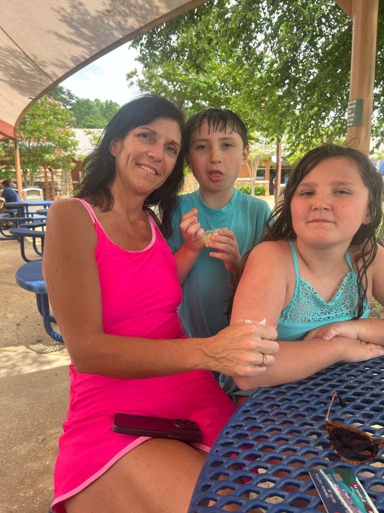 A woman and two children sitting at a table under an umbrella, enjoying a summer day at a water park.