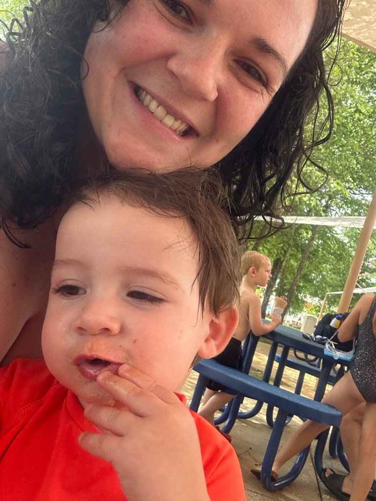A mother and her toddler enjoying a snack together, both smiling, with a backdrop of picnic tables and people at a splash pad in the background.